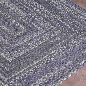 May include: Braided area rug in shades of gray and purple. The rug features a geometric pattern with a square design in the center. The rug is made of a thick, durable material, perfect for high-traffic areas.