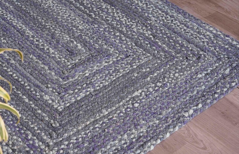 May include: Braided area rug in shades of gray and purple. The rug features a geometric pattern with a square design in the center. The rug is made of a thick, durable material, perfect for high-traffic areas.