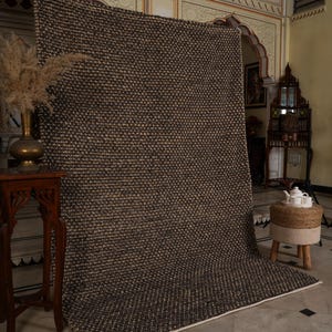May include: A large, textured area rug in shades of brown and beige. The rug has a woven appearance and a white border. It is displayed in a room with ornate architectural details and a decorative table.
