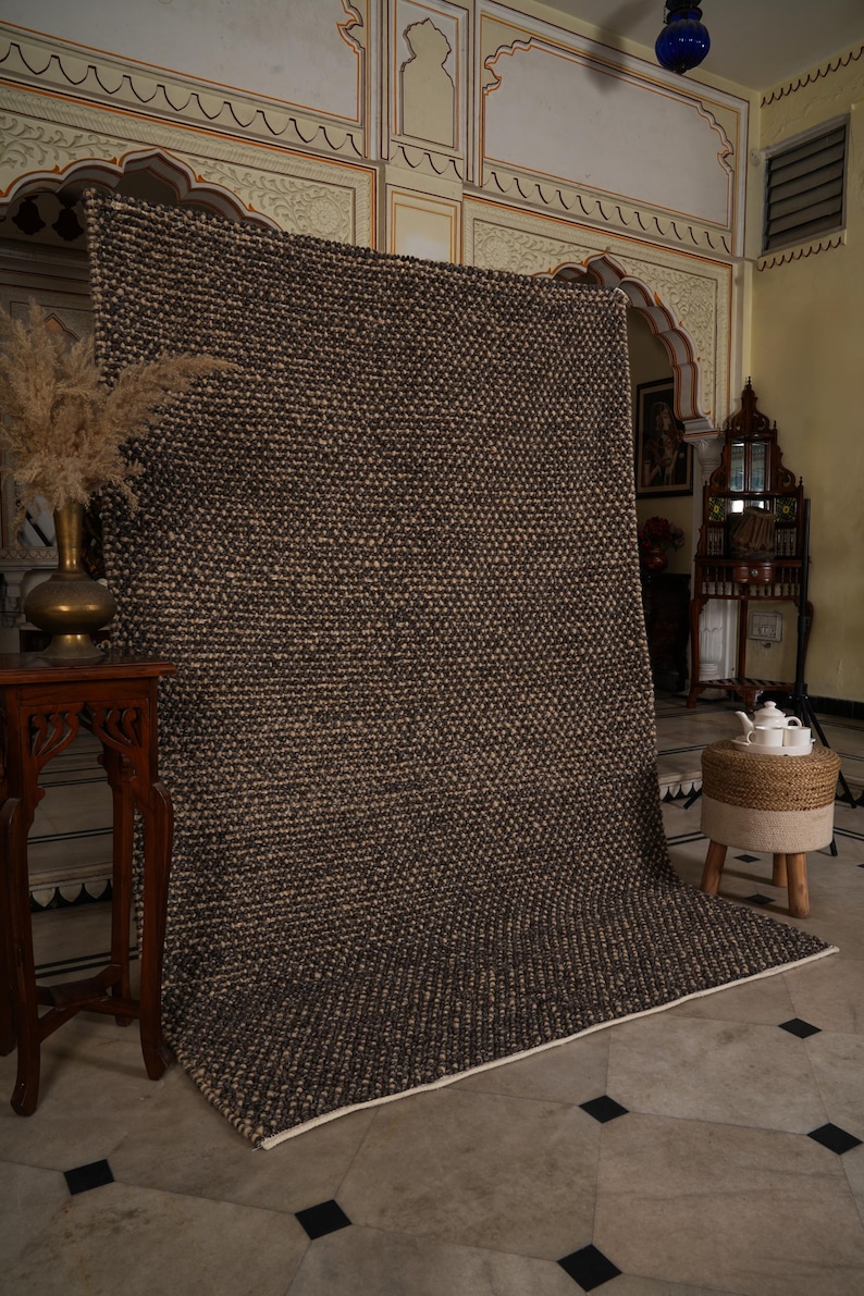 May include: A large, textured area rug in shades of brown and beige. The rug has a woven appearance and a white border. It is displayed in a room with ornate architectural details and a decorative table.