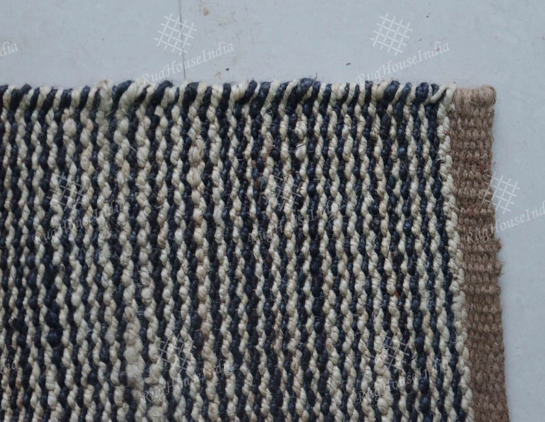 May include: A rectangular woven rug featuring a repeating vertical striped pattern in alternating dark navy blue and off-white fibers. The rug has a thick, textured braided weave construction with a solid tan border along the right edge.
