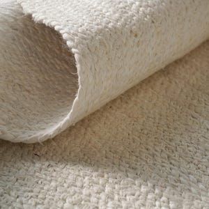 Puede incluir: Primer plano de una alfombra tejida de color blanquecino enrollada. La textura de la alfombra es visible, con un aspecto grueso y trenzado. Los bordes están cuidadosamente acabados y la alfombra parece estar hecha de fibras naturales. El color es un tono claro y neutro.