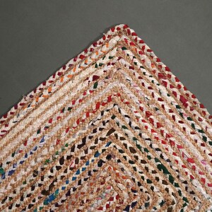 May include: Close-up of a woven rug corner. The rug features a braided design with a mix of beige, red, green, and blue threads. The rug's texture and color variations create a rustic, handmade aesthetic.