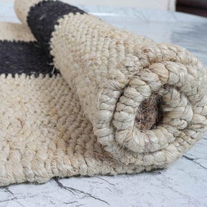 Op de afbeelding: Gerolde jute vloerkleed in natuurlijk beige met een donkergrijze streep. Het vloerkleed is gemaakt van geweven jutevezels, waardoor een textuur ontstaat. Het gerolde gedeelte toont de dikte en constructie van het vloerkleed.