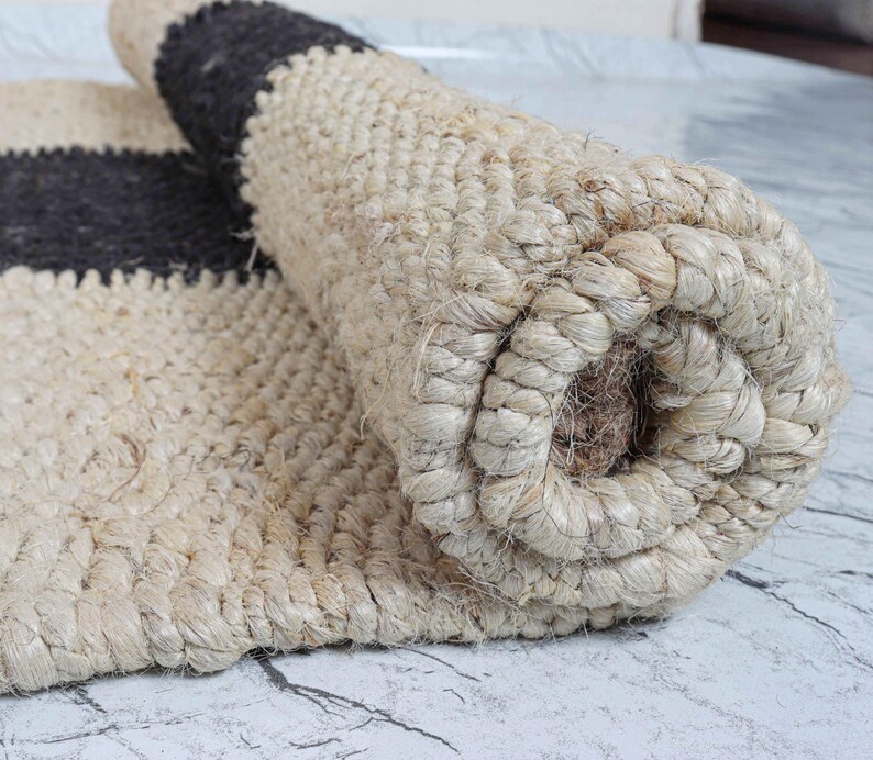 Op de afbeelding: Gerolde jute vloerkleed in natuurlijk beige met een donkergrijze streep. Het vloerkleed is gemaakt van geweven jutevezels, waardoor een textuur ontstaat. Het gerolde gedeelte toont de dikte en constructie van het vloerkleed.