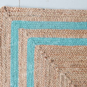 May include: A natural fiber rug with a square shape and a border of two light blue stripes. The rug is made of woven jute and has a natural, earthy tone.