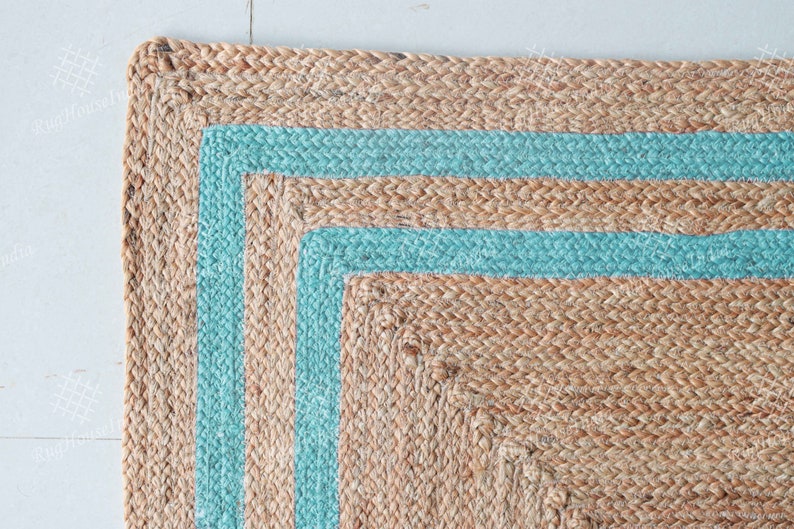 May include: A natural fiber rug with a square shape and a border of two light blue stripes. The rug is made of woven jute and has a natural, earthy tone.
