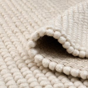 Op de afbeelding: Een dik, gebroken wit vloerkleed met een grove, handgeknoopte woltextuur en een opvallend gevlochten weefpatroon. Het kleed is op één hoek omgeslagen, waardoor de dichte lusconstructie en het stevige geweven rugmateriaal zichtbaar zijn.