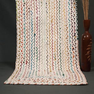 May include: A handmade, off-white crochet rug with colorful vertical stripes. The rug features a textured, open weave design with a rectangular shape. The color stripes include red, blue, yellow, and green. A brown vase with a leaf design and dried stems is in the background.