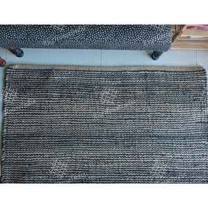 May include: A rectangular area rug featuring a woven texture with alternating horizontal stripes of dark navy blue and natural tan fibers. The rug has a thick, braided border along the top edge and a consistent, dense weave throughout the surface.