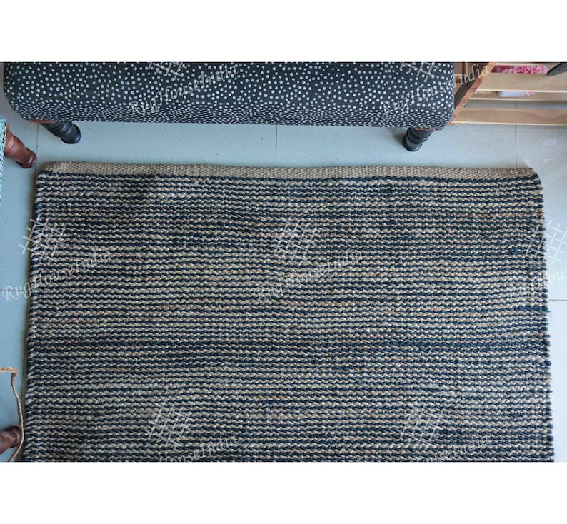 May include: A rectangular area rug featuring a woven texture with alternating horizontal stripes of dark navy blue and natural tan fibers. The rug has a thick, braided border along the top edge and a consistent, dense weave throughout the surface.