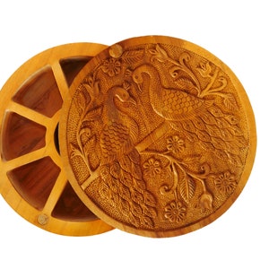 May include: A round wooden spice box with a hand-carved lid featuring two peacocks on a branch with flowers. The box has six compartments for storing spices.