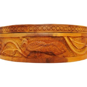 May include: A round wooden box with intricate carvings of peacocks and leaves. The box is made of light brown wood and has a lid.