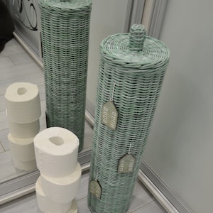May include: Two tall, cylindrical, light green woven wicker toilet paper holders with matching lids. The front holder features three small, decorative, house-shaped wooden ornaments hanging from the weave. White toilet paper rolls are stacked nearby.