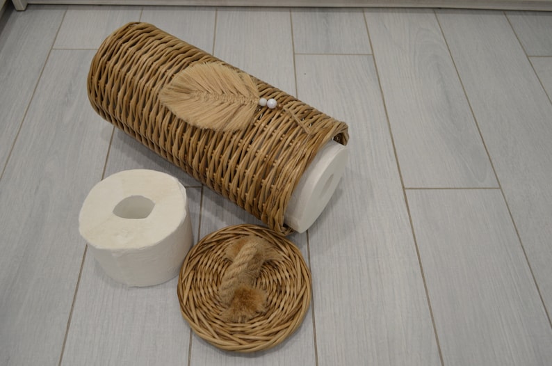 Puede incluir: Una cesta de mimbre con tapa y un rollo de papel higi&eacute;nico dentro. La cesta tiene una hoja decorativa y detalles de perlas.