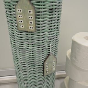 May include: A tall, cylindrical, sage green woven wicker basket featuring three hanging wooden ornaments shaped like houses. Each ornament is painted in a muted green tone with small, rectangular white windows and is suspended by a thin white string.
