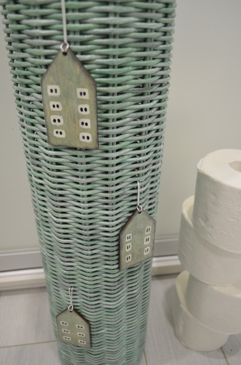 May include: A tall, cylindrical, sage green woven wicker basket featuring three hanging wooden ornaments shaped like houses. Each ornament is painted in a muted green tone with small, rectangular white windows and is suspended by a thin white string.