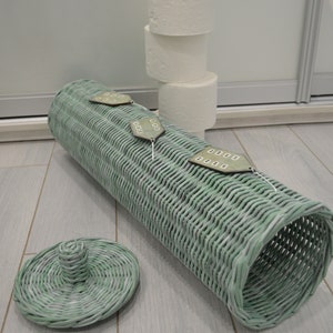 May include: A cylindrical light green woven wicker toilet paper holder with a matching circular lid. The holder features three small house-shaped wooden ornaments attached to the top, each with painted windows. Three white toilet paper rolls are stacked.