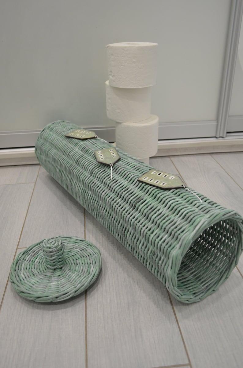 May include: A cylindrical light green woven wicker toilet paper holder with a matching circular lid. The holder features three small house-shaped wooden ornaments attached to the top, each with painted windows. Three white toilet paper rolls are stacked.