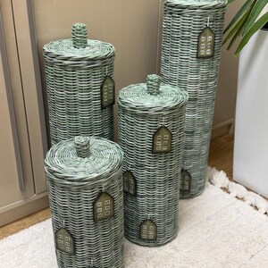 May include: Four cylindrical woven baskets in a sage green finish, each featuring a matching lid with a central knob. Each basket is adorned with small, house-shaped tags that display a grid of nine squares, attached by thin white strings.