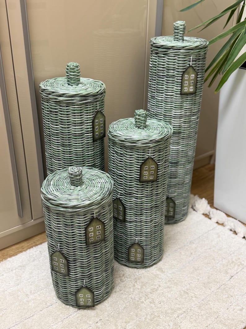 May include: Four cylindrical woven baskets in a sage green finish, each featuring a matching lid with a central knob. Each basket is adorned with small, house-shaped tags that display a grid of nine squares, attached by thin white strings.