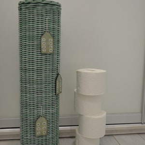 May include: A tall, cylindrical, sage green woven wicker toilet paper holder with a matching lid and a small knob handle. Three small, house-shaped wooden ornaments with white window details are hanging from the side of the wicker structure.