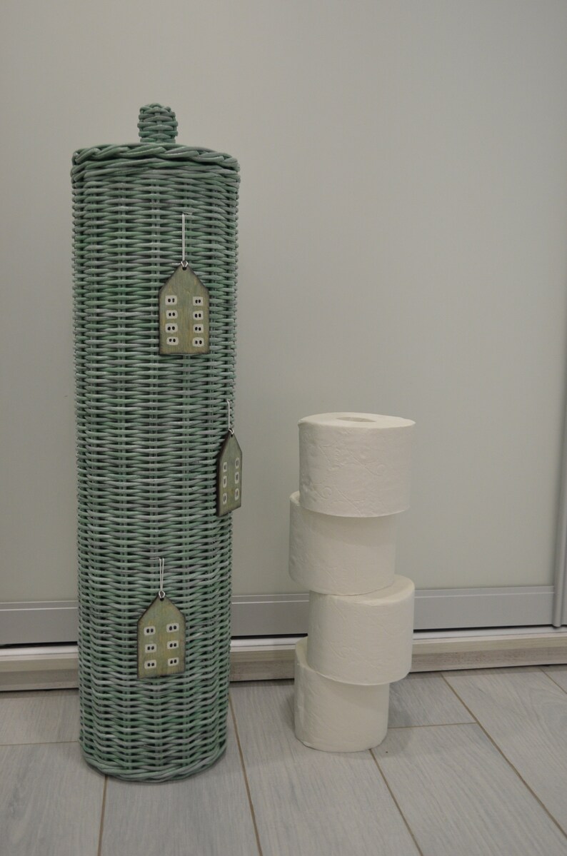 May include: A tall, cylindrical, sage green woven wicker toilet paper holder with a matching lid and a small knob handle. Three small, house-shaped wooden ornaments with white window details are hanging from the side of the wicker structure.