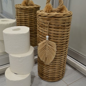 Puede incluir: Dos cestas de mimbre con tapas y asas de cuerda. Una cesta tiene una pluma marr&oacute;n y una cuenta blanca colgando de ella. Las cestas est&aacute;n sobre un suelo de baldosas de color gris claro. Hay cuatro rollos de papel higi&eacute;nico blanco apilados junto a las cestas.
