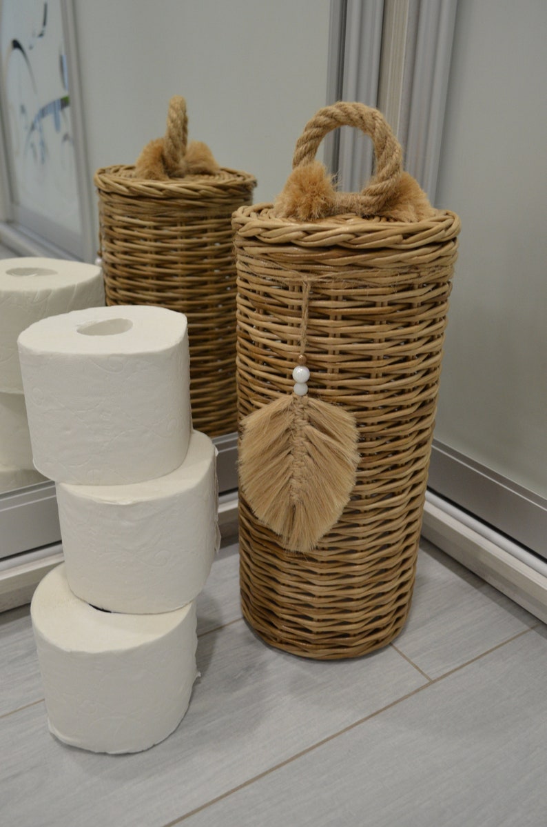 Puede incluir: Dos cestas de mimbre con tapas y asas de cuerda. Una cesta tiene una pluma marr&oacute;n y una cuenta blanca colgando de ella. Las cestas est&aacute;n sobre un suelo de baldosas de color gris claro. Hay cuatro rollos de papel higi&eacute;nico blanco apilados junto a las cestas.