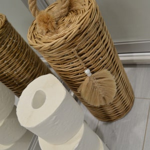 Puede incluir: Un porta rollo de papel higi&eacute;nico de mimbre con asa de cuerda y borla marr&oacute;n. Tres rollos de papel higi&eacute;nico est&aacute;n apilados debajo del soporte.