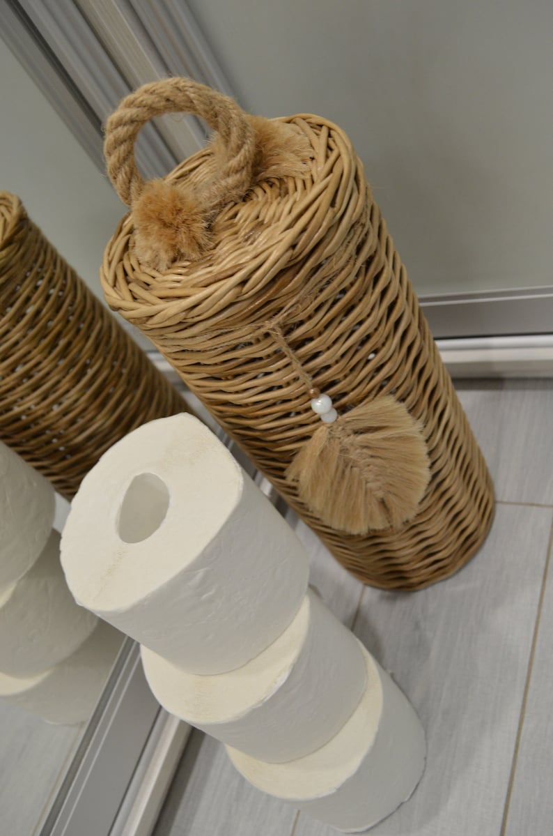Puede incluir: Un porta rollo de papel higi&eacute;nico de mimbre con asa de cuerda y borla marr&oacute;n. Tres rollos de papel higi&eacute;nico est&aacute;n apilados debajo del soporte.