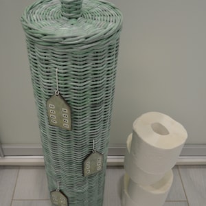 May include: A tall, cylindrical, light green woven wicker toilet paper holder with a matching lid and a knob handle. Three small, flat, house-shaped wooden ornaments with white window details are attached to the front. Three white toilet paper rolls.