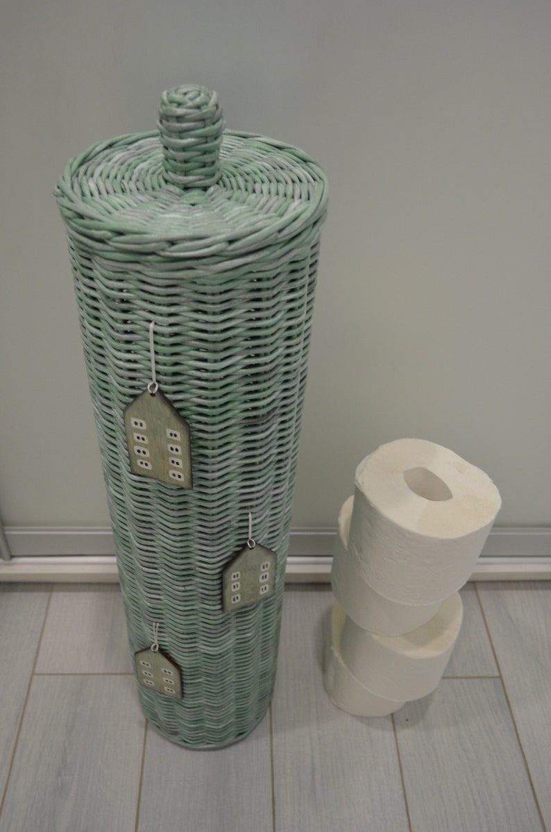 May include: A tall, cylindrical, light green woven wicker toilet paper holder with a matching lid and a knob handle. Three small, flat, house-shaped wooden ornaments with white window details are attached to the front. Three white toilet paper rolls.