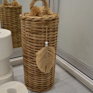 Puede incluir: Un porta rollo de papel higi&eacute;nico de mimbre tejido con un asa de cuerda y un borla de plumas marrones. El soporte est&aacute; sobre un suelo de madera clara.