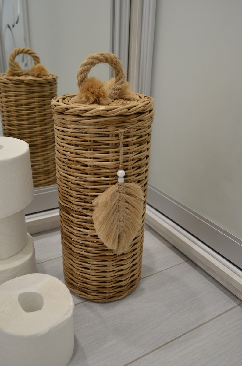 Puede incluir: Un porta rollo de papel higi&eacute;nico de mimbre tejido con un asa de cuerda y un borla de plumas marrones. El soporte est&aacute; sobre un suelo de madera clara.