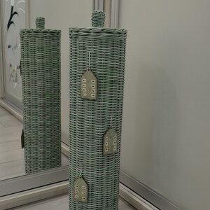 May include: A tall, cylindrical, sage green woven wicker toilet paper holder with a matching lid. Three small, flat, wooden house-shaped ornaments with white window details are hanging vertically down the front of the woven structure.