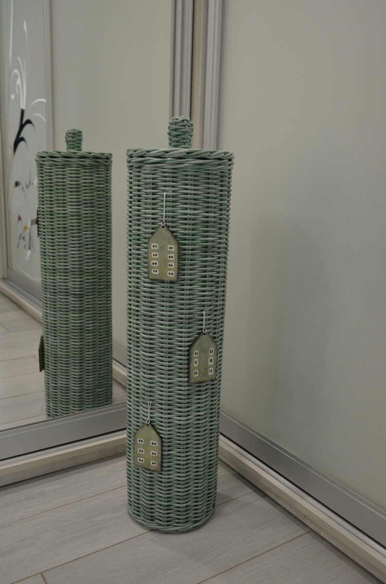 May include: A tall, cylindrical, sage green woven wicker toilet paper holder with a matching lid. Three small, flat, wooden house-shaped ornaments with white window details are hanging vertically down the front of the woven structure.