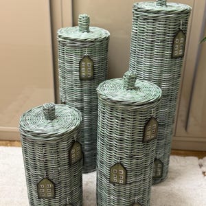 May include: Four cylindrical sage green woven wicker storage baskets with lids. Each basket features a small knob on the lid and is decorated with multiple house-shaped tags hanging from white strings, each displaying a grid of small squares.