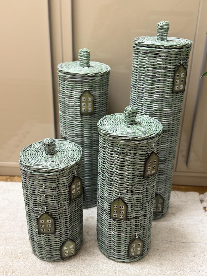May include: Four cylindrical sage green woven wicker storage baskets with lids. Each basket features a small knob on the lid and is decorated with multiple house-shaped tags hanging from white strings, each displaying a grid of small squares.