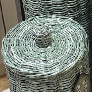 May include: A cylindrical woven basket with a matching lid, featuring a light sage green finish with subtle grey undertones. The lid has a central woven knob handle, and a small house-shaped tag hangs from the side of the basket structure.