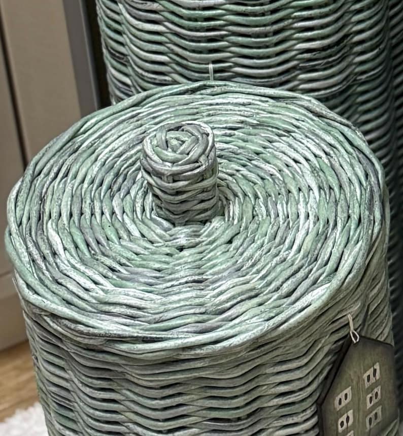 May include: A cylindrical woven basket with a matching lid, featuring a light sage green finish with subtle grey undertones. The lid has a central woven knob handle, and a small house-shaped tag hangs from the side of the basket structure.