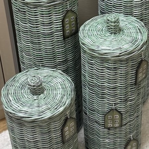 May include: A set of four cylindrical sage green woven wicker containers with matching lids, each featuring a small knob handle on top. Every container is adorned with a house-shaped tag displaying a grid of small white squares on a dark background.