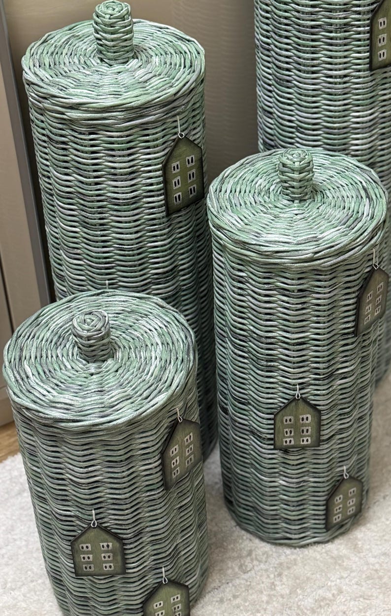 May include: A set of four cylindrical sage green woven wicker containers with matching lids, each featuring a small knob handle on top. Every container is adorned with a house-shaped tag displaying a grid of small white squares on a dark background.