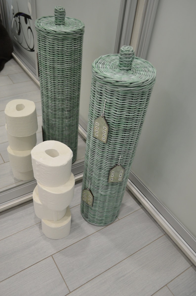 May include: Two tall, cylindrical, light green woven wicker toilet paper holders with matching lids. The front holder features three small, decorative, house-shaped wooden ornaments hanging from the weave. White toilet paper rolls are stacked nearby.