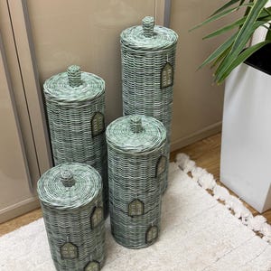 May include: A set of four cylindrical woven containers in a pale sage green color. Each container features a matching lid with a small central knob and a decorative house-shaped tag attached to the side with a thin string, creating a uniform look.