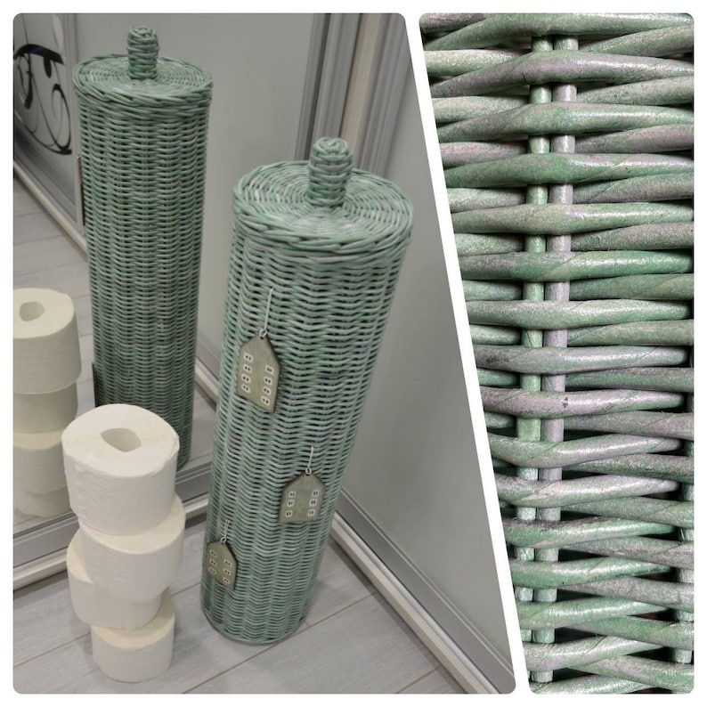 May include: Two tall, cylindrical, sage green woven wicker toilet paper holders with matching lids and small knobs. One holder features three hanging wooden house-shaped ornaments. A stack of four white toilet paper rolls sits beside them.