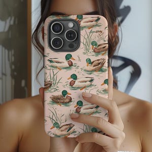 May include: A phone case featuring a repeating pattern of ducks swimming in a pond. The ducks have green heads, white and brown bodies, and are set against a light pink background. Green reeds and water details complete the design.