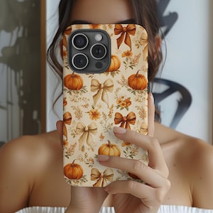 May include: A phone case featuring a fall-themed pattern. The design includes orange pumpkins, tan bows, and floral accents on a cream background. The case has a camera cutout and is held in front of a person.