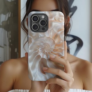 Op de afbeelding: Een witte en beige iPhone-hoes met een swirlend schelpmotief. De hoes heeft een verhoogde camerabult met drie zwarte lenzen.