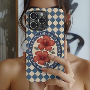May include: A phone case with a blue and cream diamond pattern. The center features a floral design with two red hibiscus flowers. The case has a camera cutout and is held in front of a person.